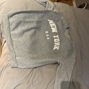 Grå sweatshirt med New York-tryck - Grå sweatshirt med vit text 'NEW YORK U.S.A.' på bröstet. Klassisk rund halsringning och långa ärmar. Mjuk och skön, perfekt för chill dagar eller när du vill ha en enkel streetstyle-look.
