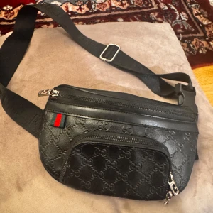 Svart  magväska i skinn - Snygg svart magväska präglat skinn med GG-mönster över hela väskan. Väskan har två dragkedjefack, justerbart band och klassisk grön-röd detalj. Perfekt för att bära det viktigaste och ge din outfit en lyxig touch.