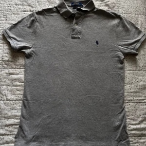 Grå pikétröja från Polo Ralph Lauren - Snygg grå pikétröja från Polo Ralph Lauren i slim fit-modell. Klassisk krage med två knappar och broderad marinblå logga på bröstet. Tillverkad i mjuk bomull, perfekt för en clean och stilren look.