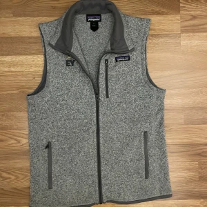 Grå fleeceväst från Patagonia - Säljer en grå fleeceväst från Patagonia i storlek S. Västen har hel dragkedja framtill, tre fickor med dragkedja och en hög krage. Materialet är 100% återvunnen polyester och den har en snygg, melerad look med mörkare grå detaljer vid krage och fickor.
