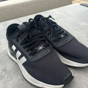 Svarta Adidas sneakers med vita ränder - Snygga svarta Adidas sneakers med klassiska vita ränder på sidorna och svart/vit sula. Ovandelen är i textil med detaljer i syntet och snörning framtill. Perfekta för dig som gillar en sportig och stilren look.