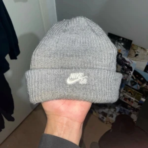 Grå stickad mössa från Nike SB - Snygg grå stickad mössa från Nike SB med uppvikt kant och broderad vit logga framtill. Perfekt för att hålla värmen och addera streetstyle till din look. Enkel design som passar till de flesta outfits.