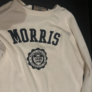Vit stickad tröja från Morris - Vit stickad tröja från Morris med mörkblå elbow patches och stort tryck framtill med logga och texten 'MORRIS'. Tröjan har ribbade muddar och rund hals. Perfekt för dig som gillar klassisk och stilren preppy-look.