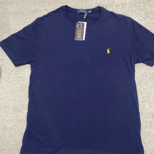Marinblå t-shirt från Polo Ralph Lauren - Snygg marinblå t-shirt från Polo Ralph Lauren med klassisk gul broderad logga på bröstet. Rund hals och korta ärmar. Tillverkad i 100% mjuk bomull för skön känsla. Perfekt för dig som gillar stilrena och ikoniska plagg.