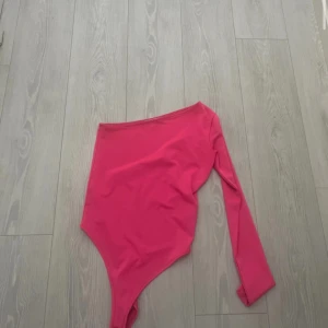 NEON Rosa one shoulder topp - Snygg rosa topp med one shoulder-design och lång ärm på ena sidan. Tight passform som framhäver siluetten och ger en trendig look. Perfekt att styla med jeans eller kjol till fest.