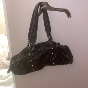 Svart axelväska med nitar och vita sömmar - Cool svart axelväska i skinnimitation med breda handtag, dekorativa nitar och kontrasterande vita sömmar. Väskan har dragkedja upptill, små sidofickor och snygga metalldetaljer. 