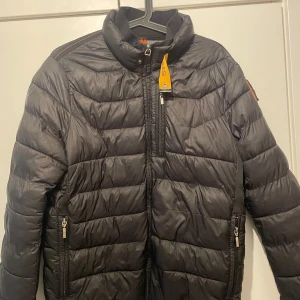 Svart lättviktsdunjacka Parajumpers M - Svart dunjacka från Parajumpers i storlek M. Jackan är superlätt, quiltad och har dragkedjor på fickorna samt en hög krage. Klassisk patch på ärmen och snygga detaljer i gult vid halsen. Perfekt för kyliga dagar när du vill hålla stilen. Dock har jackan små hål men dom är bra tejpade och dragkedjan är lite sönder men går och stänga.