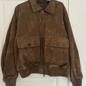 Brun mockajacka från Gant - Säljer en brun mockajacka från Gant med klassisk krage, två stora fickor framtill med knappar och grov dragkedja. Jackan har ribbade muddar och en schysst loose passform. Insidan är fodrad med slätt skinn för extra premiumkänsla. Perfekt för dig som gillar stilren och tidlös look. Den är helt oanvänd och funkar som unisex såklart! Ordinarie pris 12 000kr