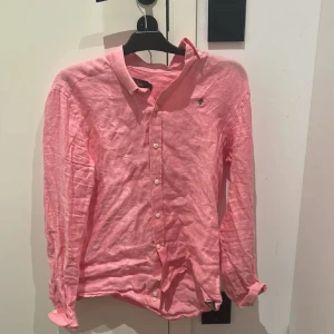 Rosa linneskjorta från Morris - Snygg rosa skjorta från Morris i 100% linne med klassisk passform. Skjortan har långa ärmar, knappar framtill och en liten broderad logga på bröstet. Perfekt för dig som gillar en fräsch och avslappnad stil.