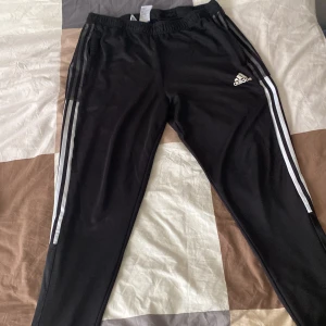 Svarta Adidas träningsbyxor med ränder - Svarta träningsbyxor från Adidas med klassiska vita ränder längs sidorna och Adidas-logga på låret. Byxorna har elastisk midja och är gjorda i ett mjukt, syntetiskt material som passar perfekt för sport eller chill. Snygg och sportig look.