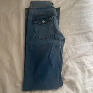 Blå bootcut jeans från Nelly - Snygga blå jeans från Nelly med bootcut passform och hög midja. Jeansen har råa kanter nertill, stora framfickor och dubbla knappar i midjan. Perfekta för dig som gillar en retro vibe och vill ha ett par jeans som sticker ut lite extra.