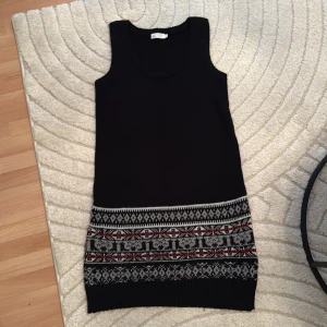 Stickad svart klänning från att. - Svart ärmlös stickad klänning från att. i storlek M. Klänningen har ett brett mönstrat parti nertill i grått, vitt och rött med klassiskt nordiskt inspirerat mönster. Rund halsringning och ribbade kanter. Perfekt för lager-på-lager-stil under kallare månader.