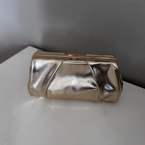 Guldig kuvertväska med kedja  - Snygg kuvertväska i  guld med mjuk, veckad design och guldfärgad kedja. Väskan har hård ram och stilren låsdetalj. Perfekt att bära i handen eller över axeln för en extra touch till din outfit.