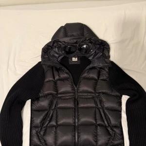 Svart dunväst med huva från C.P. Company - Säljer en svart dunväst från C.P. Company med ikonisk goggle-huva och dragkedja framtill. Västen har quiltad design och stickade svarta ärmar, vilket ger en unik look. Perfekt för dig som vill sticka ut med streetwear-vibe och funktionella detaljer.