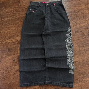 Svart lång jeanskjol från JNCO - Grym svart långkjol i jeans från JNCO med fet grå drak-inspirerad print längs ena sidan och broderad krona på bakfickan. Klassisk femficksmodell med coola detaljer och bred passform. Perfekt för dig som vill sticka ut med streetstyle-vibe.