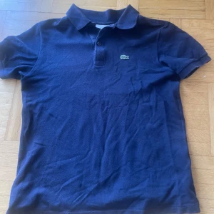 Marinblå pikétröja från Lacoste - Snygg marinblå pikétröja från Lacoste med klassisk krage och knappslå framtill. Ikoniska krokodilloggan broderad på bröstet. Tillverkad i mjuk bomull som känns skön mot huden. Perfekt för en stilren och sportig look.