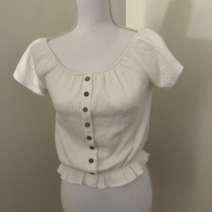 Vit kortärmad blus med knappar - Charmig vit blus med korta ärmar och knappar framtill. Den har en söt volangdetalj vid nederkanten och en rundad halsringning. Perfekt för en somrig look!