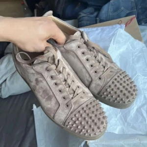 Louboutin skor - Säljer ett par snygga beige sneakers från Christian Louboutin. Skorna har en unik design med nitar på tån och snörning framtill. De har den ikoniska röda sulan som är typisk för märket. De är mycket bra skick. Hör av dig vid frågor eller prisförslag!