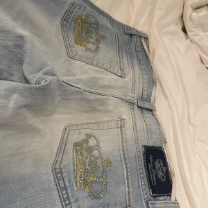 Ljusblå jeans från Victoria Beckham for Rock & Republic - Snygga ljusblå jeans med broderade guldkronor på bakfickorna. De har en klassisk femficksdesign och en knappgylf. Perfekta för en stilren look. Skriv för mer information (fickorna har tappat få stenar)