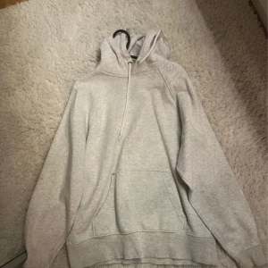 Grå hoodie - Säljer en klassisk grå hoodie med luva och känguruficka framtill. Perfekt för en avslappnad stil. Den har långa ärmar och ribbade muddar vid ärmslut och nederkant.