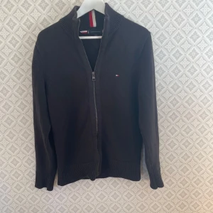 Svart cardigan  från Tommy Hilfiger - Säljer en stilren svart kofta från Tommy Hilfiger med dragkedja och ribbade detaljer. Den har en liten logotyp på bröstet och röd-vit randig detalj vid kragen. Perfekt för en klassisk look.