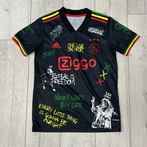 Ajax fotbollströja med Bob Marley-tema - Snygg svart fotbollströja från Adidas med Bob Marley-tema. Tröjan har korta ärmar med rasta-färger på kanterna och flera grafiska tryck och citat i olika färger. Perfekt för fotbollsfans som älskar musik och stil.