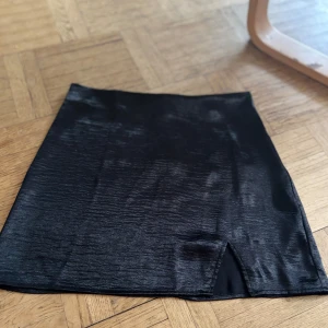 Svart kjol från Gina Tricot - Snygg svart kjol från Gina Tricot i en glansig finish. Kjolen har en liten slits framtill och en dragkedja bak för enkel påklädning. Perfekt för en stilren look.