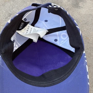 Blå keps från Boss - Snygg blå keps från Boss med ett iögonfallande vitt mönster av diagonala ränder och text. Perfekt för att ge din outfit en sportig touch. Kepsen har en klassisk passform och justerbar rem baktill.