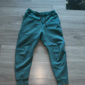 Turkosa mjukisbyxor från Nike - Säljer ett par turkosa mjukisbyxor från Nike med snörning i midjan och elastiska muddar vid bensluten. Byxorna har en avslappnad passform och är perfekta för en bekväm stil. Nike-loggan är tryckt på framsidan.