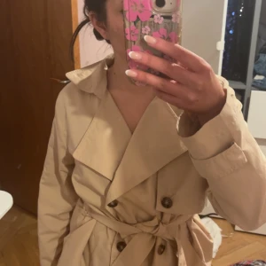 Beige trenchcoat med bälte från vero moda - Snygg beige trenchcoat med knappar och bälte i midjan. Jackan har en klassisk krage och långa ärmar, perfekt för vår och höst. Den har en stilren design som passar till många olika outfits.
