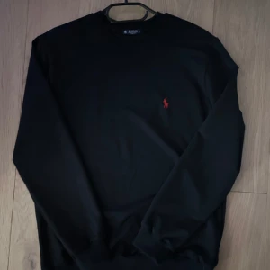 Svart tröja från Ralph Lauren - Säljer en stilren svart tröja från Ralph Lauren med det klassiska röda logotypen på bröstet. Tröjan har långa ärmar och en normal passform. Perfekt för en avslappnad och snygg look.