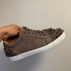 Christian Louboutin - Snygga bruna sneakers från Christian Louboutin med nitar på tå och häl. Skorna har en vit sula och är tillverkade i mocka med snörning framtill. Perfekta för att ge din stil en edgy touch.
