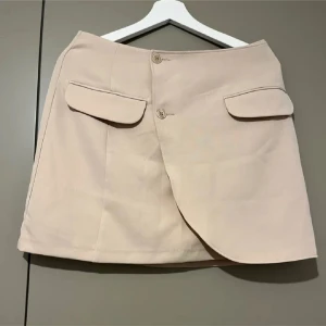 Beige kortkjol med knappar - Snygg beige kortkjol med omlott-design och knappar framtill. Kjolen har två dekorativa fickor och en stilren look. Perfekt för en chic och modern stil.