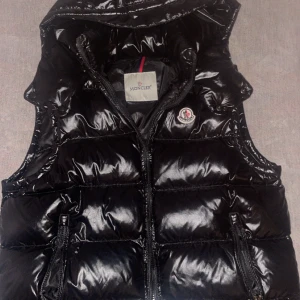 Moncler väst svart - Snygg svart dunväst från Moncler med glansig yta och dragkedja framtill. Västen har en broderad logotyp på bröstet och praktiska fickor med dragkedja. Perfekt för kyligare dagar.