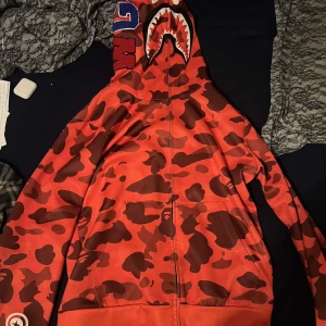 Röd bape hoodie  - Säljer en röd bape  hoodie med camo och hajmotiv på huvan. Hoodien har dragkedja och broderade detaljer på sidan av huvan. Perfekt för en unik streetwear-look. Priser kan diskuteras 