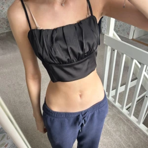 Svart satinlinnetopp - Snygg svart satinlinnetopp med smala axelband och rynkad detalj framtill. Perfekt för en stilren och festlig look.