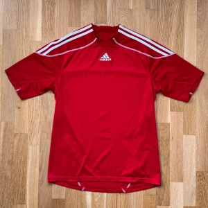 Adidas Röd T-Shirt - Adidas funktions/tränings/fotbolls T-Shirt i färgen röd. Storlek: M, Armhåla till Armhåla: 50 cm, Längd: ~70 cm fram och ~73 cm, Halsöppning till Armöppning: 43 cm.  Material: 100% Polyester.