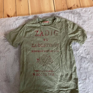 Olivgrön t-shirt från Zadig & Voltaire - Säljer en olivgrön t-shirt från Zadig & Voltaire med tryck i rött på framsidan. T-shirten har en klassisk rund hals och korta ärmar. Perfekt för en avslappnad stil. Skicka ett meddelande vid intresse!