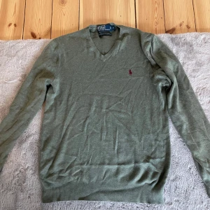 Grön tröja från Polo Ralph Lauren - Säljer en stilren grön tröja från Polo Ralph Lauren i 100% pima bomull. Tröjan har en v-ringad design och långa ärmar med ribbade muddar. Perfekt för en avslappnad och elegant look.