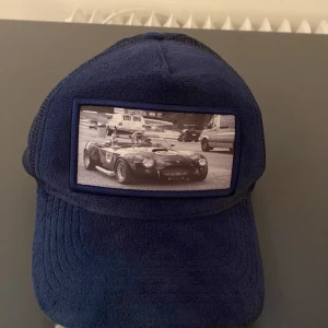 Blå keps från Emilio Pendenza - Snygg blå keps från Emilio Pendenza med ett coolt bilmotiv på framsidan. Kepsen har ett nätmaterial på baksidan för extra ventilation. Perfekt för en avslappnad stil.