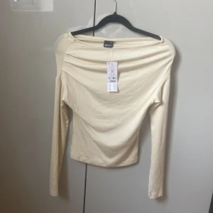 Beige asymmetrisk topp från Gina Tricot - Säljer en stilren beige asymmetrisk topp från Gina . Toppen har långa ärmar och en offshoulder-design som ger en trendig look. Perfekt för att lyfta vilken outfit som helst. Passar bra till både jeans och kjol. 🌟(aldrig använt)
