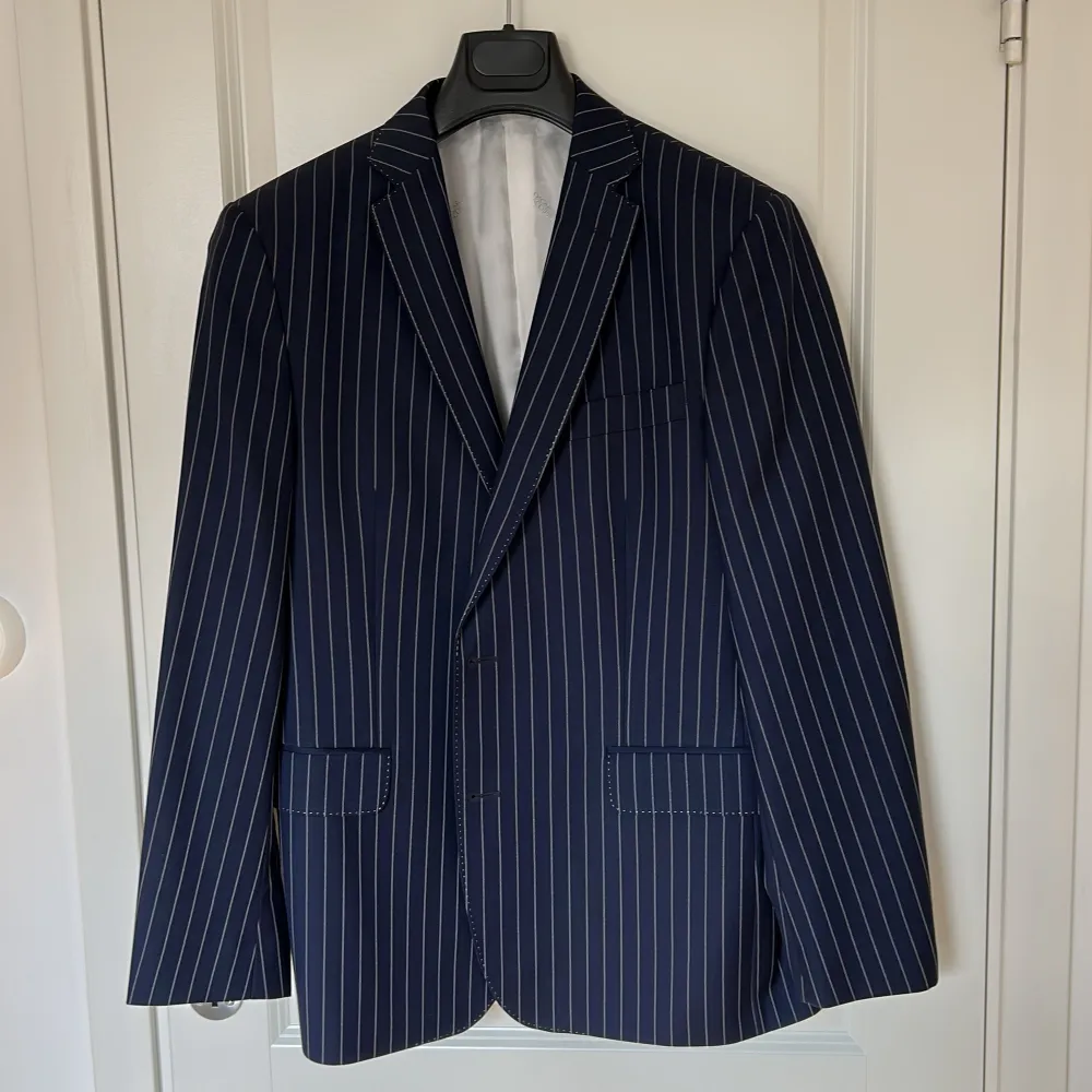 Snygg mörkblå kavaj från Oscar Jacobson med vita ränder och eleganta detaljer. Kavajen har en klassisk passform med knappar framtill och vita armbågslappar som ger en stilfull touch. Perfekt för en sofistikerad look.. Puvut & Bleiserit.