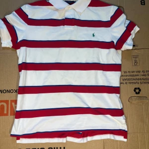 Randig pikétröja från Ralph Lauren - Säljer en snygg randig pikétröja från Ralph Lauren i vitt, rött och blått. Tröjan har korta ärmar och en klassisk krage med knappar. Perfekt för en stilren och avslappnad look. 🏷️OBSS!! Tröjan är custom fit så har ni några frågor om passform fråga!!! 