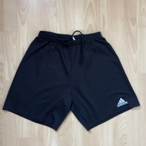 Svarta träningsshorts från Adidas - Snygga svarta träningsshorts från Adidas med elastisk midja och snörning. Perfekta för gymmet eller löprundan. Adidas-loggan i vitt på benet ger en sportig touch.
