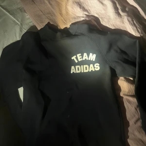 Svart långärmad tröja från Adidas - Säljer en svart långärmad tröja från Adidas med texten 'TEAM ADIDAS' i vitt på framsidan. Perfekt för träning eller en avslappnad dag. Tröjan har en klassisk passform och är tillverkad i ett mjukt material.