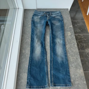 Lågmidjade bootcut jeans - Säljer ett par snygga blå bootcut jeans med slitna detaljer. De har en klassisk femficksdesign och knappar fram. Perfekta för en avslappnad stil. Midjemått: 38cm midjehöjd:19 cm innerbenslängd: 76cm
