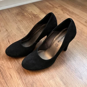 Svarta pumps / klackar från Tamaris - Snygga svarta pumps från Tamaris i mocka med rund tå och klack. Perfekta för en elegant look. Skorna har en klassisk design som passar till många tillfällen.