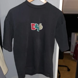 Svart t-shirt från District Fortysix - Säljer en svart t-shirt från District Fortysix med ett färgglatt D46-tryck på framsidan. T-shirten är kortärmad och tillverkad i bomull, perfekt för en avslappnad stil.(Små skador)