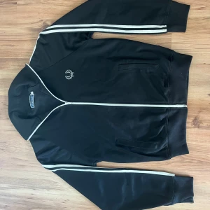 Svart track jacket från Fred Perry - Säljer en svart track jacket från Fred Perry med vita ränder längs ärmarna och broderad logga på bröstet. Jackan har dragkedja och ribbade muddar vid ärmslut och nederkant. Perfekt för en sportig look! (Pris kan diskuteras vid upphämtning)