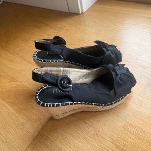 Svarta kilklackar med rosett - Snygga svarta kilklackar  med öppen tå och dekorativ rosett framtill. Skorna har en flätad sula som ger en somrig känsla och en justerbar rem med knappdetalj vid hälen.